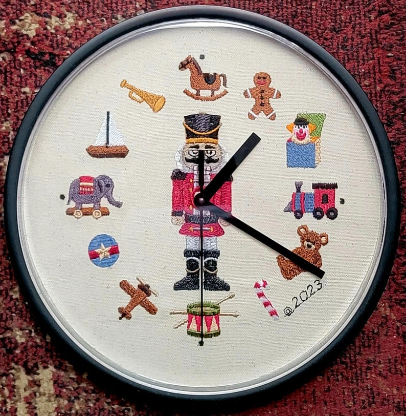 May include: A white clock face with a black frame and black hands. The clock face is embroidered with various toys, including a nutcracker, a rocking horse, a toy train, a teddy bear, a toy drum, a toy aeroplane, a toy boat, a toy elephant, a gingerbread man, a toy trumpet, a toy box, a candy cane, and a red and blue ball. The clock face also includes the text "@2023".