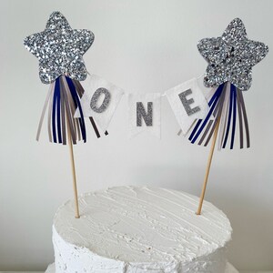 Puede incluir: Un pastel blanco de dos pisos con una estrella plateada de purpurina y una pancarta blanca que dice "ONE" en la parte superior. La pancarta está decorada con serpentinas azules, grises y marrones.