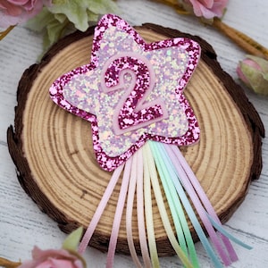 Könnte beinhalten: Ein sternförmiges Abzeichen mit einem dunkelrosa Glitzerrand und einer hellen, irisierenden Glitzermitte. Die Zahl 2 ist mittig in dunkelrosa Glitzer aufgebracht. Ein Bündel dünner, pastellfarbener Satinbänder in Rosa, Gelb, Grün und Blau hängt herab.