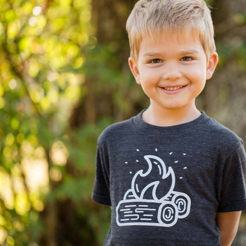May include: A young boy wearing a dark grey t-shirt with a white graphic of a campfire on it. The boy is smiling and looking at the camera.