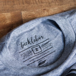 May include: A gray t-shirt with a black tag that says "tacklebox" and includes washing instructions. The tag also includes the size "2" and the text "75% cotton 25% rayon".