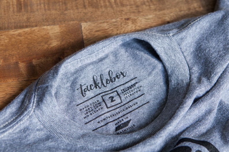 May include: A gray t-shirt with a black tag that says "tacklebox" and includes washing instructions. The tag also includes the size "2" and the text "75% cotton 25% rayon".