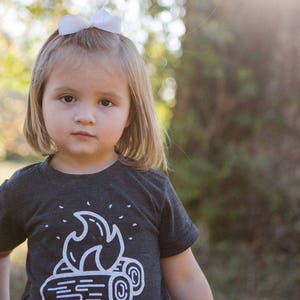 May include: A young girl with blonde hair wearing a black t-shirt with a white campfire graphic. She is wearing a white bow in her hair and has a serious expression on her face.