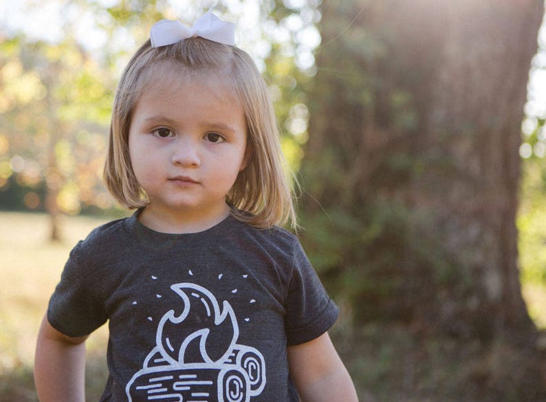 May include: A young girl with blonde hair wearing a black t-shirt with a white campfire graphic. She is wearing a white bow in her hair and has a serious expression on her face.