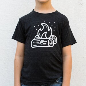 May include: A dark grey t-shirt with a white graphic of a campfire with flames and logs.