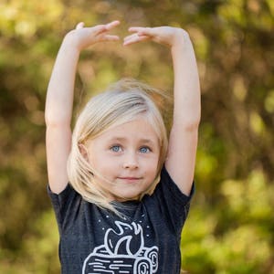 May include: A young girl with blonde hair is wearing a dark grey t-shirt with a white graphic of a campfire on it. She is standing in front of a blurred background of green trees.