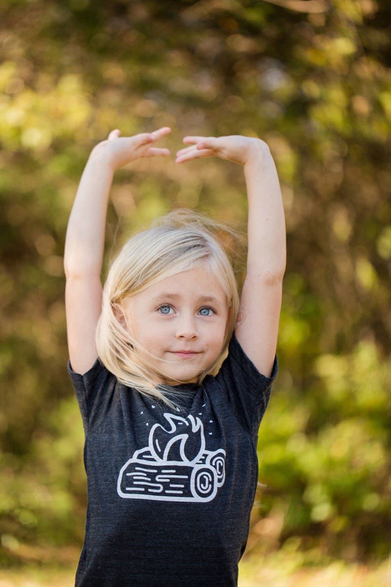 May include: A young girl with blonde hair is wearing a dark grey t-shirt with a white graphic of a campfire on it. She is standing in front of a blurred background of green trees.