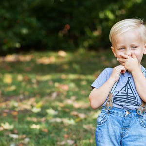 May include: A young boy with blonde hair is wearing a light blue t-shirt with a sailboat graphic and blue jeans with suspenders. He is smiling and covering his mouth with his hand.