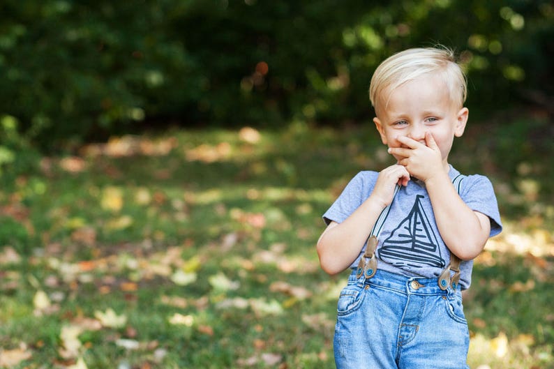 May include: A young boy with blonde hair is wearing a light blue t-shirt with a sailboat graphic and blue jeans with suspenders. He is smiling and covering his mouth with his hand.