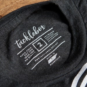 May include: A black t-shirt with a white tag that says "tacklebox" and includes care instructions, size 2, website address, and "Made in Nashville".