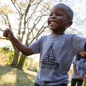 May include: A young boy wearing a grey t-shirt with a black sailboat graphic is laughing and raising his arms in the air. He is outdoors in a grassy area with trees in the background.