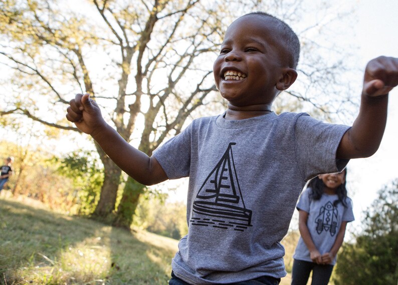 May include: A young boy wearing a grey t-shirt with a black sailboat graphic is laughing and raising his arms in the air. He is outdoors in a grassy area with trees in the background.