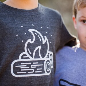 May include: A dark gray t-shirt with a white graphic of a campfire. The graphic features a stylized flame over two logs.