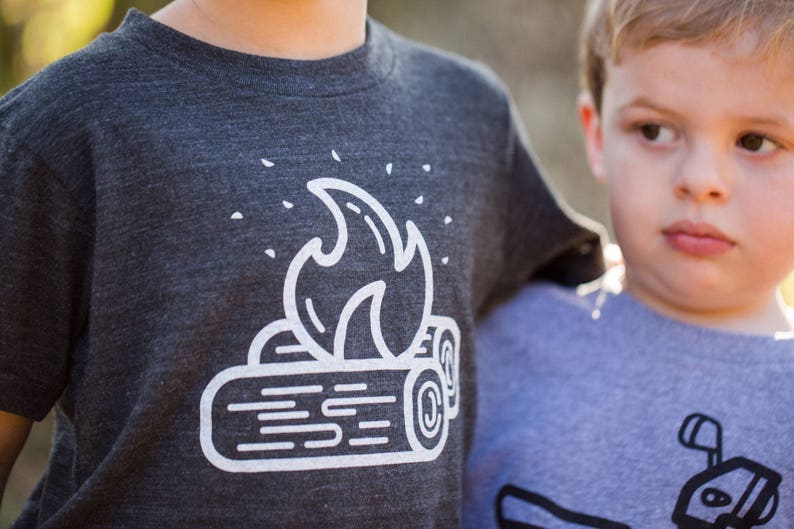May include: A dark gray t-shirt with a white graphic of a campfire. The graphic features a stylized flame over two logs.