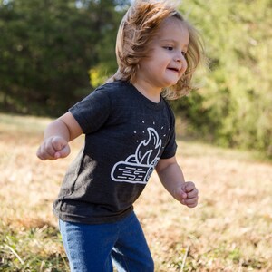 May include: A young child wearing a dark grey t-shirt with a white graphic of a campfire on it. The child is running through a field of tall grass.