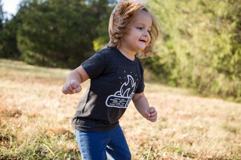 May include: A young child wearing a dark grey t-shirt with a white graphic of a campfire on it. The child is running through a field of tall grass.