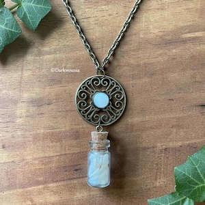 Può includere: Una collana con catena color bronzo e un ciondolo circolare in filigrana con un cabochon bianco centrale. Una piccola fiala di vetro con tappo di sughero, riempita di frammenti bianchi simili a petali, pende dalla parte inferiore.