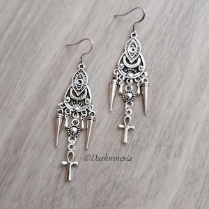 Peut inclure: Une paire de boucles d'oreilles pendantes en argent avec un design complexe représentant un œil, une lune croissante et un symbole ankh. Les boucles d'oreilles sont rehaussées de petites perles et de pointes argentées.