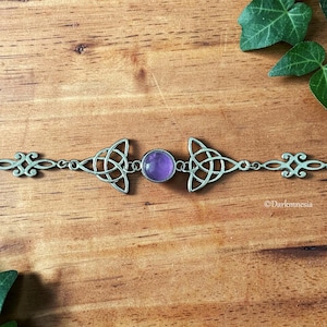 May include: A silver bracelet with Celtic knot motifs and a central, round, purple gemstone. The bracelet is displayed on a wooden surface. The design incorporates symmetrical patterns and decorative elements. The bracelet is approximately 18 cm in length.