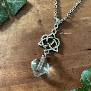 Könnte beinhalten: Silberfarbene Halskette mit einem keltischen Knoten-Anhänger und einem klaren, facettierten Glasfläschchen. Das Fläschchen hängt unter dem Knoten, die Kette ist vor einem Holzgrund zu sehen. Ein einzigartiges Schmuckstück.