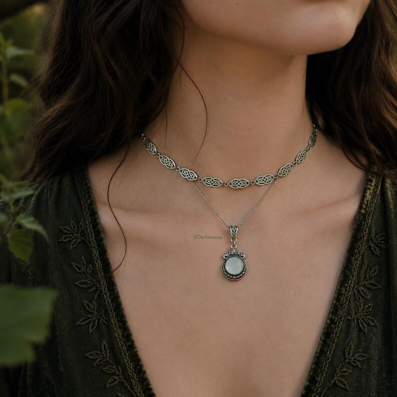 May include: A silver-coloured necklace set. The top necklace is a choker with an openwork Celtic knot design, and the lower necklace has a pendant with a round, light-coloured stone. The necklaces are layered.
