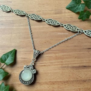 May include: A silver necklace with a Celtic knot design. The necklace has a double-strand upper section and a pendant with a round, pale green stone. The pendant is framed by a decorative silver setting. The background is a wooden surface with green leaves.