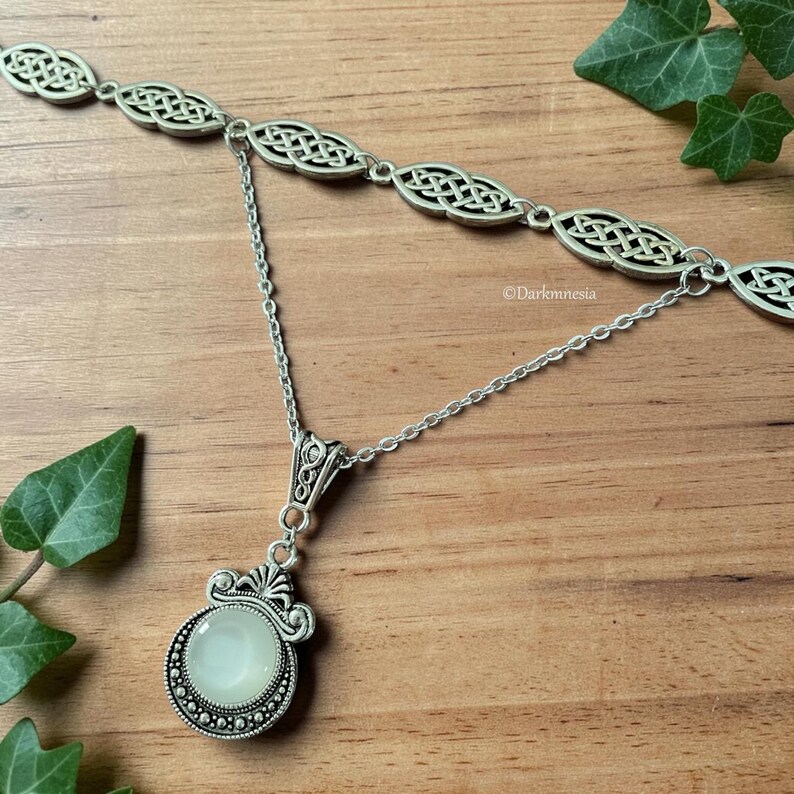 May include: A silver necklace with a Celtic knot design. The necklace has a double-strand upper section and a pendant with a round, pale green stone. The pendant is framed by a decorative silver setting. The background is a wooden surface with green leaves.