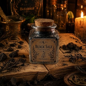 Könnte beinhalten: Ein quadratisches Glasgefäß gefüllt mit schwarzem Salz, verschlossen mit einem Korkstopfen. Das Etikett zeigt einen Halbmond, Farnillustrationen und den Text: Black Salt, Protection & Banishing, Darkmnesia. Das Glas ist klar und durchsichtig.