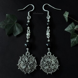 Peut inclure: Une paire de boucles d'oreilles pendantes argentées. Chaque boucle d'oreille comprend un crochet, une chaîne de perles noires et d'éléments argentés, et un pendentif circulaire avec un motif d'étoile. Les boucles sont sur fond sombre avec des feuilles vertes.