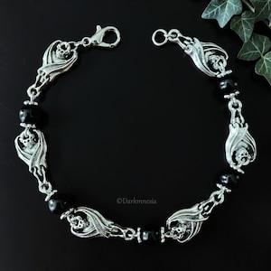 Op de afbeelding: Zilverkleurige armband met vleermuis- en doodshoofdbedels afgewisseld met zwarte kralen. De armband heeft een karabijnsluiting en wordt op een donkere achtergrond getoond. Het ontwerp heeft een gotische of alternatieve esthetiek.