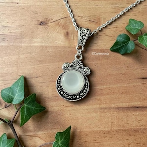 May include: A silver pendant necklace featuring a round, off-white stone. The pendant has an ornate, filigree design with a beaded border. The silver chain rests on a wooden surface with green ivy. The pendant is a classic design.
