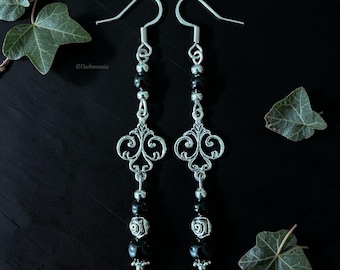May include: A pair of silver-toned dangle earrings with black beads and ornate filigree designs. The earrings have a hook closure and are displayed against a dark background with green leaves. The earrings have a gothic or vintage aesthetic.
