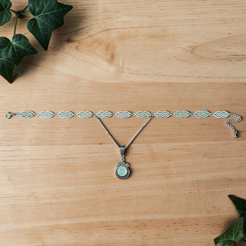 May include: A silver necklace and bracelet set with a Celtic knot design. The necklace features a pendant with a light blue stone. The jewellery is displayed on a wooden surface, with ivy leaves in the background.
