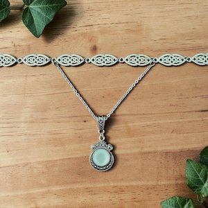 May include: A silver necklace and choker set. The choker has a Celtic knot design. The pendant necklace features a round, pale green stone with a decorative silver frame. The jewellery is displayed on a wooden surface.