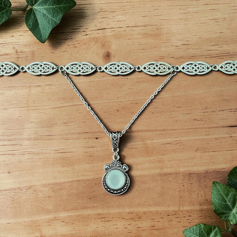 May include: A silver necklace and choker set. The choker has a Celtic knot design. The pendant necklace features a round, pale green stone with a decorative silver frame. The jewellery is displayed on a wooden surface.