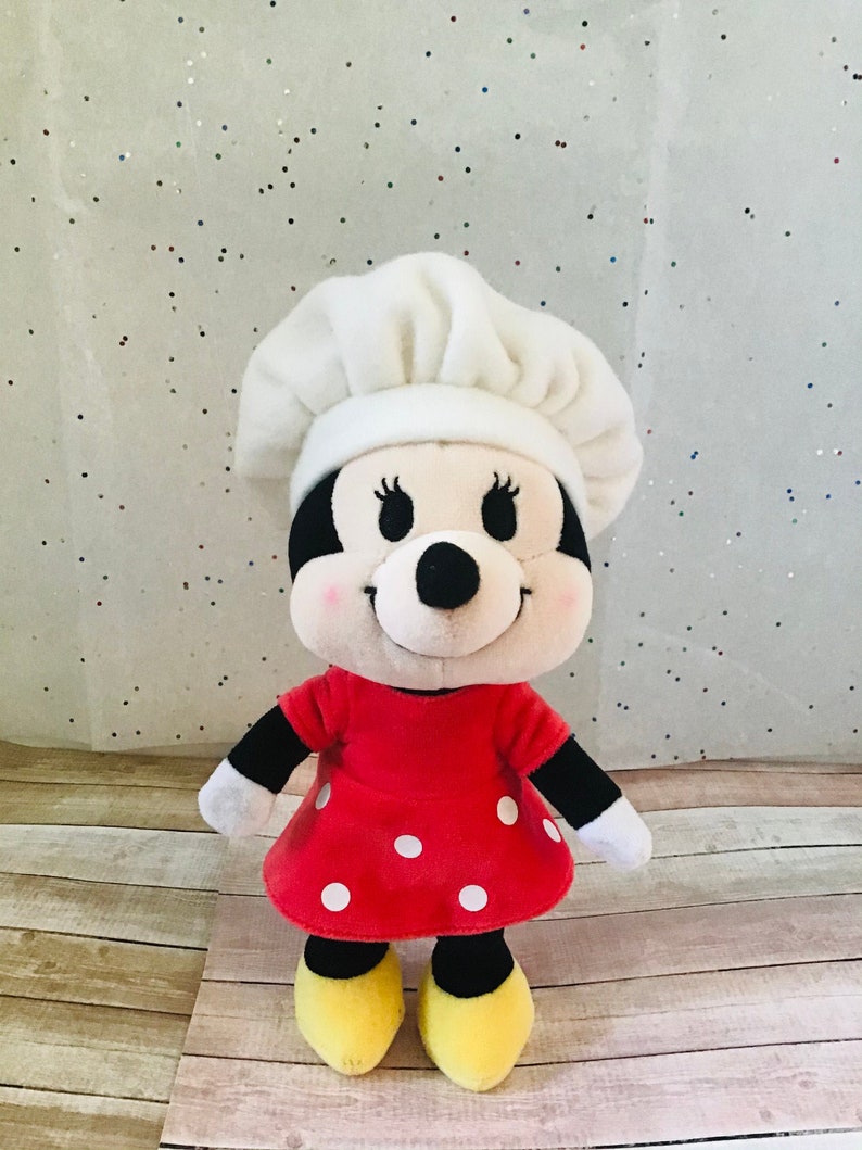 Pu&ograve; includere: Un giocattolo di peluche di un personaggio dei cartoni animati che indossa un cappello da chef bianco e un vestito rosso con pois bianchi. Il giocattolo ha occhi neri, un naso nero e un blush rosa sulle guance.
