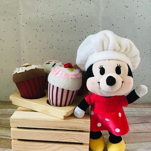 Pu&ograve; includere: Un giocattolo di peluche a forma di topo con un cappello da chef e un vestito rosso a pois bianchi. Il topo &egrave; in piedi davanti a una cassa di legno con tre cupcake.