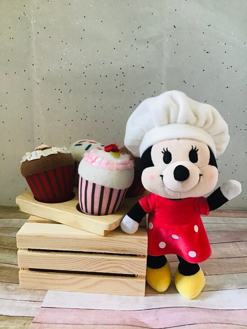 Pu&ograve; includere: Un giocattolo di peluche a forma di topo con un cappello da chef e un vestito rosso a pois bianchi. Il topo &egrave; in piedi davanti a una cassa di legno con tre cupcake.