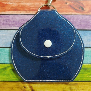 Może przedstawiać: Niebieski brelok z brokatem, z małą kieszonką i srebrnym zapięciem. Brelok ma biały guzik z przodu.