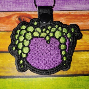 May include: A purple and green embroidered keychain shaped like a cauldron with bubbling green liquid. The keychain has a black leather strap with a metal ring.