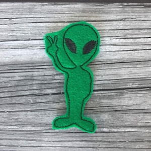 Puede incluir: Un extraterrestre de fieltro verde con ojos negros y un gesto de la mano de la paz. El extraterrestre est&aacute; delineado en negro y est&aacute; de pie. El fondo es una superficie de madera gris.