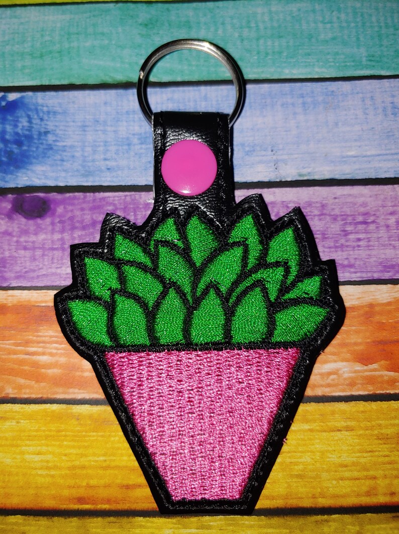 Puede incluir: Un llavero de cuero negro con un bot&oacute;n rosa, que representa una planta suculenta verde en una maceta rosa.
