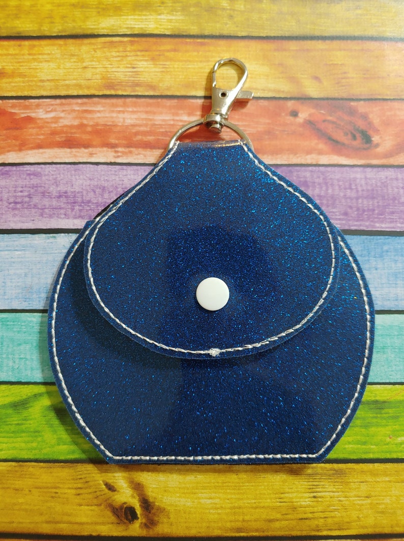 Może przedstawiać: Niebieski brelok z brokatem, z małą kieszonką i srebrnym zapięciem. Brelok ma biały guzik z przodu.