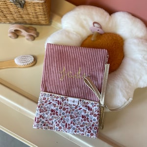 Peut inclure: Un carnet rectangulaire avec une couverture en velours côtelé rose poudré portant le prénom Jady brodé en fil doré. La partie inférieure présente un imprimé floral dans des tons de rose, rouge et gris, avec un ruban marque-page doré sur le côté.