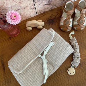 Peut inclure: Un tapis à langer en tissu beige à texture gaufrée avec un ruban blanc, une voiture jouet en bois, une paire de sandales en cuir marron, une attache-sucette en tissu beige et un dahlia rose dans un vase en verre sur une surface en bois.