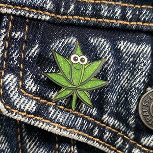 May include: A green enamel pin in the shape of a cannabis leaf with a smiling face and googly eyes. The pin is set against a dark blue denim jacket.