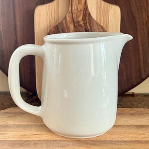 May include: A white ceramic pitcher with a curved handle and a pouring spout. The pitcher is set against a backdrop of wooden cutting boards in various shades of brown, creating a rustic aesthetic. The pitcher is likely used for serving beverages.