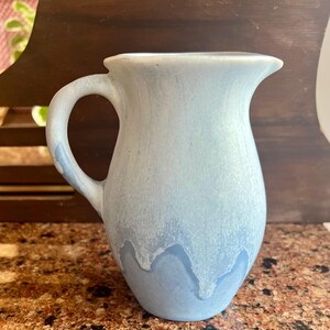 May include: A light blue ceramic pitcher with a handle and a spout. The pitcher has a gradient effect, transitioning from a lighter blue at the top to a darker blue at the bottom. The handle is curved and attached to the side of the pitcher.