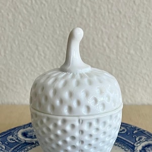 May include: A white, lidded jar with a textured, dimpled surface, resembling a strawberry. The jar has a curved stem-like handle on top. It sits on a blue and white patterned plate, creating a charming, vintage aesthetic.