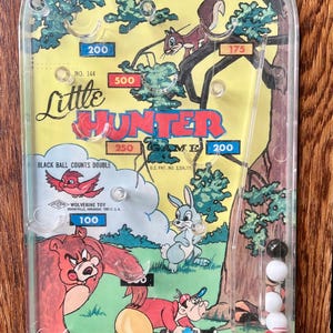 May include: Vintage "Little Hunter" pinball game with colorful illustrations of forest animals and score values. The game features a clear plastic cover, a plunger, and a series of holes for the steel balls to fall into.